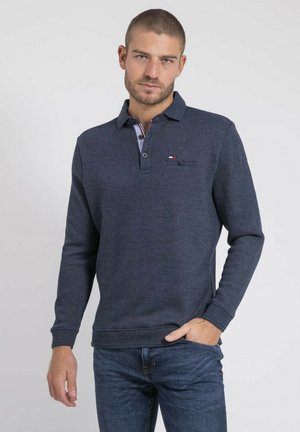 Homme aux cheveux courts portant un polo à manches longues bleu marine et un jean bleu, debout avec une main dans la poche sur un fond uni.