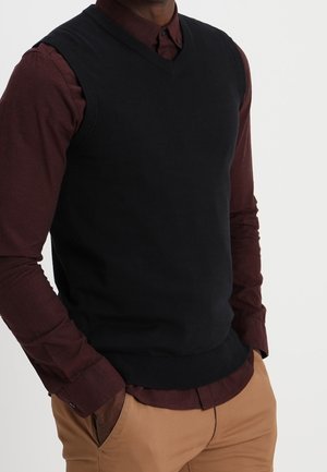 Homme portant un pull sans manches noir par-dessus une chemise à boutons bordeaux, avec un pantalon beige, les mains dans les poches.
