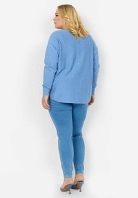 Šviesiai mėlynas tamprių rankovių marškinėlis su mygtukais nugaros pusėje, derinamas su pritaikytomis mėlynomis džinsais ir mėlynais aukštakulniais mules, prieš baltą fono.