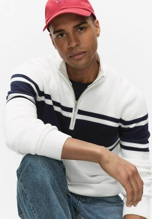 Mężczyzna w czerwonej czapce i białym swetrze z granatowymi paskami, siedzący z jedną nogą podciągniętą do góry, patrzący w kamerę na białym tle.