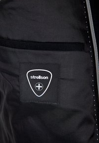 Wnętrze czarnej kurtki z tkaniny z zamkniętą kieszenią oraz metką z logo w kształcie tarczy, podkreślającą markę "Strellson" w srebrnym kolorze.