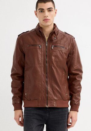 Joven mirando al frente, vistiendo una chaqueta de cuero marrón con cremallera, bolsillos en el pecho con cremallera y puños y dobladillo acanalados.