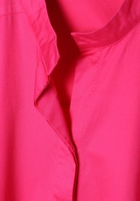 Chemise rose vif avec un col à encolure pliée et une texture lisse, légèrement brillante. Le tissu semble frais et léger.