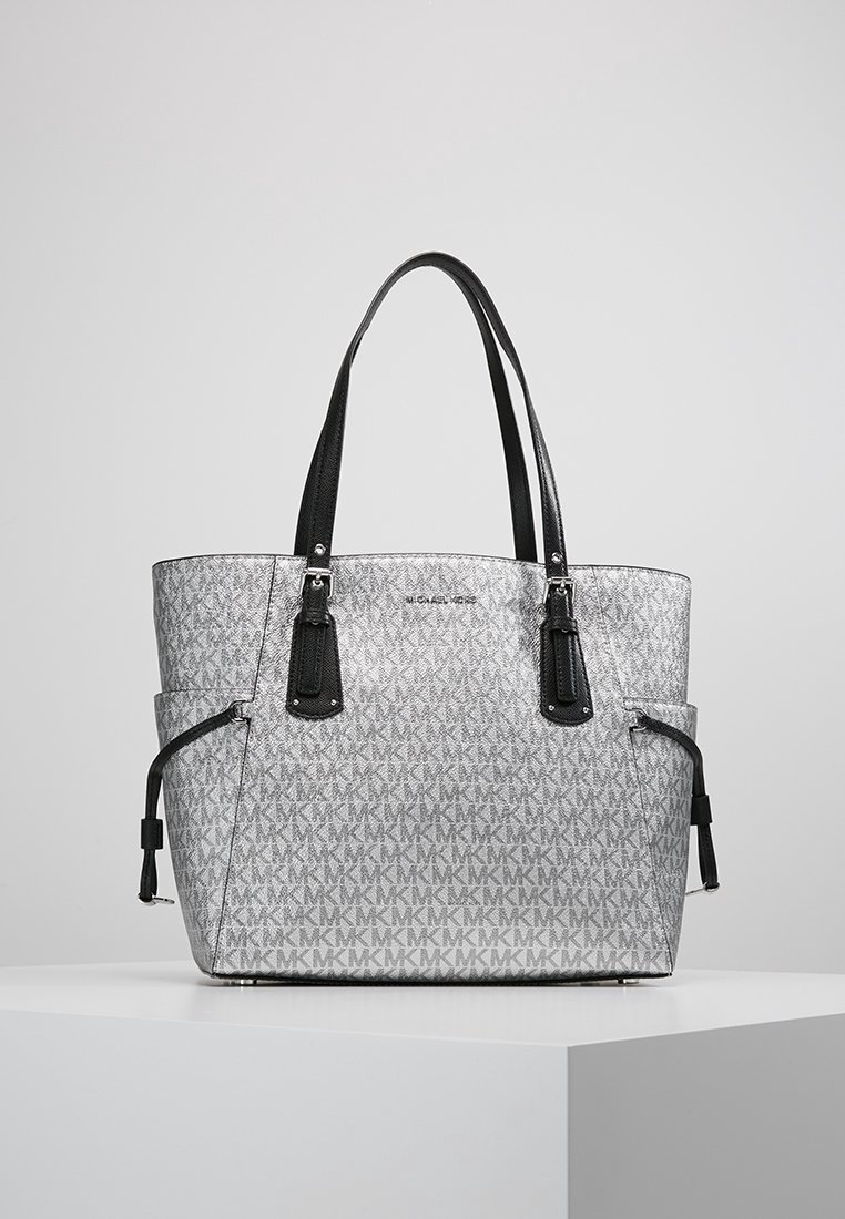 Sac fourre-tout gris avec motif répétitif "MK", poignées noires, poches latérales et logo "Michael Kors" à l'avant, posé sur une surface blanche.