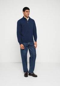Camisola de malha azul-marinho com gola alta e fecho de correr, combinada com jeans de ganga azul claro e sapatos castanhos de vestir. Design simples e ajustado.
