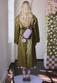 Zöld, fényes ballonkabát barna pántos táskával és kör alakú, mintás pénztárcával. Magassarkú szandálok, virágos ruha alatta, hullámos haj.