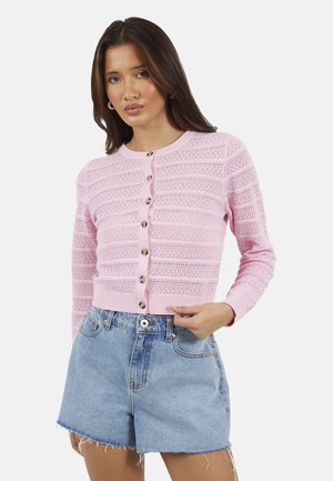 Kobieta ubrana w różowy zapinany na guziki sweter z dzianiny z poziomymi wzorami oraz jasnoniebieskie dżinsowe szorty z wysokim stanem i postrzępionymi brzegami.