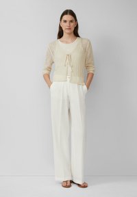 Cardigan beige tricoté avec un nœud à l’avant, superposé sur un débardeur léger, associé à un pantalon blanc large et des sandales marron.