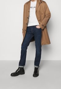 Brun trenchcoat över vit t-shirt med svart text, mörkblå jeans och svarta snörstövlar med strukturerad sula.