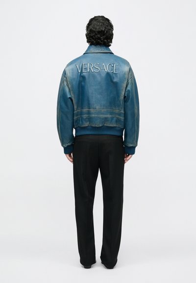 Άνδρας με ξεθωριασμένο μπλε δερμάτινο μπουφάν με μεγάλο λογότυπο "VERSACE" στην πλάτη, μαύρο παντελόνι, στέκεται κοιτώντας προς λευκό φόντο.