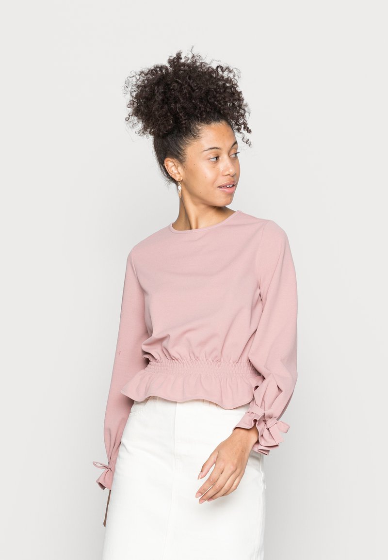 Pink long-sleeve top with a smocked waist, ruffled hem, and bow details at the sleeves, made from a soft fabric. Worn with a light-colored skirt.
