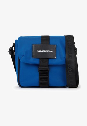 Bolso bandolera de nailon azul con solapa frontal, correa negra con logo y forma rectangular. Cierre asegurado con una hebilla negra.