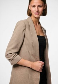 Blazer beige con una finitura leggermente testurizzata. Caratterizzato da un colletto a scialle, maniche a tre quarti e tasche frontali. Indossato sopra un top nero.