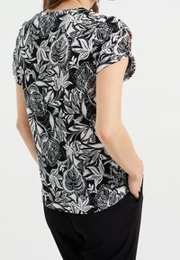 Černobílá květinová blůza s krátkými rukávy, zakřiveným lemem a lehkým materiálem s jemnými vzory listů. V kombinaci s černými kalhotami.
