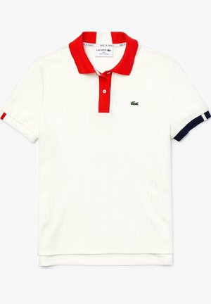 Personne en jupe plissée blanche, polo blanc avec col rouge, chaussettes blanches et baskets blanches, se penchant en avant avec les bras tendus.