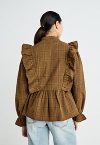 Blusa a scacchi marrone con spalle e schiena arricciate, vita a peplo e maniche ampia. Realizzata in tessuto texture, con colletto alto.
