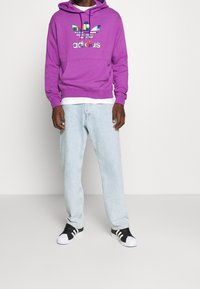 Lila huvtröja med färgglad Adidas-logotyp, kombinerad med ljusblå jeans och svart-vita sneakers. Slät textur.