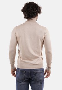 Maglione beige a collo alto lavorato a maglia con polsini e orlo a coste, design aderente, texture liscia e dettagli posteriori senza cuciture. Indossato con jeans blu.