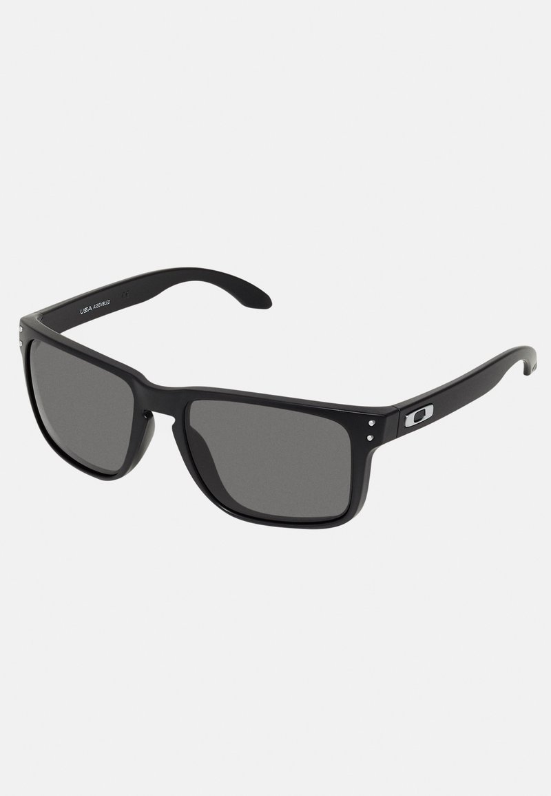 Ochelari de soare negri cu un cadru din plastic gros, lentile gri închis și accente metalice subtile pe brațe. Formă pătrată cu textură netedă.