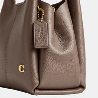 Taupe színű, szemcsés bőr kézitáska aranyszínű fémrészekkel, és egy bőr márkajelzéses címkével, amely golyóláncra van fűzve.