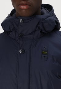 Navy blue puffer jacket with a rounded hood, button closure, and a yellow logo tag on the chest. Smooth, insulated fabric texture.