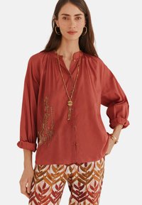 Rotes Hemd mit Knopfleiste, gerafftem Ausschnitt, langen Ärmeln und goldener Blumenstickerei. Kombiniert mit gemusterten Hosen mit Blätterdesigns.