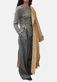 Grijze geribbelde lange mouwtop met knoopaccenten, gecombineerd met losse grijze broek en een tan lange jas van een zacht suede-achtig materiaal.