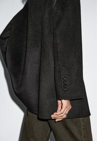 Czarny wełniany żakiet o luźnym kroju, z klapami i szczegółami z czterema guzikami na mankietach. Połączony z oliwkowymi spodniami o teksturowanym wykończeniu.