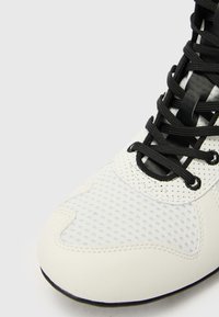 Scarpa sportiva bianca con pannelli in rete, lacci neri e superficie testurizzata. Design con punta arrotondata e suola piatta per stabilità.