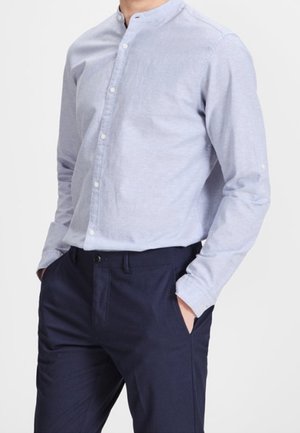 Homme portant une chemise bleu clair boutonnée avec un col mandarin et un pantalon bleu marine foncé, les mains dans les poches.