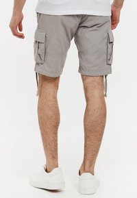 Grijze cargoshorts met opgerolde zoom, voorzien van zijzakken en strikken. Draagt witte sneakers, tegen een effen achtergrond.