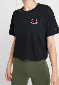 Černé zkrácené tričko vyrobené z měkké tkaniny, s výrazným vyšívaným logem v červené a bílé barvě na hrudi a krátkými rukávy.