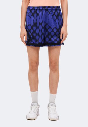 Personne portant un short de sport bleu et noir à motifs, des chaussettes blanches et des baskets blanches, debout sur un fond uni.