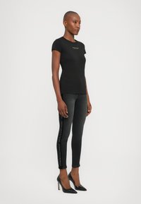Vrouw in een aansluitend zwart T-shirt met logo en zwarte skinny jeans met zijbranding, gecombineerd met zwarte hoge hakken, staande in een zijaanzicht.