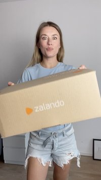 Caja de cartón de Zalando con una textura marrón clara y un logo blanco, sostenida por una persona con una camisa azul y pantalones cortos de mezclilla desgastados.