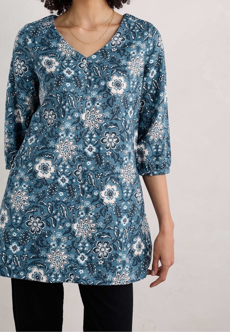 Femme portant une tunique bleue à motif floral avec des manches trois-quarts et un col en V, associée à un pantalon noir et des colliers délicats en or.