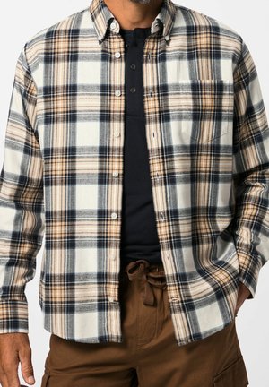 Homme portant une chemise à carreaux beige, blanche et noire boutonnée sur un t-shirt henley noir, associée à un pantalon marron avec cordon.