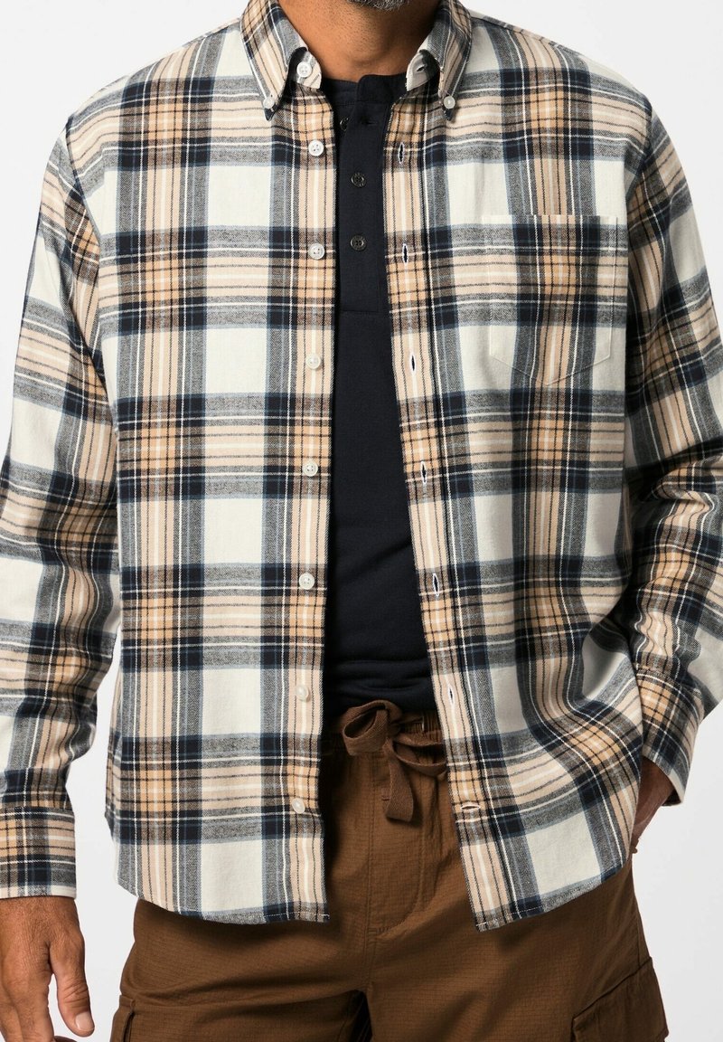 Homme portant une chemise à carreaux beige, blanche et noire boutonnée sur un t-shirt henley noir, associée à un pantalon marron avec cordon.