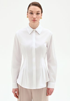 Donna che indossa una maglia bianca aderente a maniche lunghe con pieghe e pantaloni beige, in piedi davanti a uno sfondo chiaro uniforme.