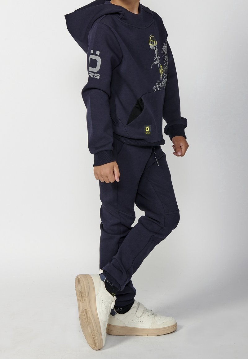 Enfant portant un sweat à capuche bleu marine et un pantalon assorti avec des baskets blanches, debout de profil sur un fond clair uni.