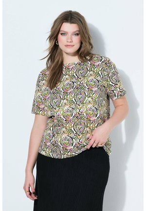 Femme aux cheveux bruns ondulés portant un haut à manches courtes multicolore à motif cachemire et une jupe plissée noire, posant devant un mur blanc.