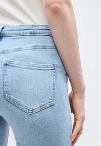 Κοντινό πλάνο του χεριού ενός ατόμου που ακουμπάει δίπλα σε τζιν τζιν με ανοιχτό μπλε χρώμα, πίσω τσέπη και ορατή ραφή.