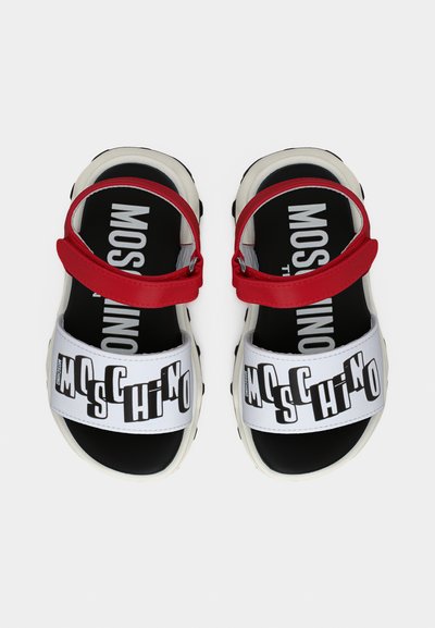Λευκά και μαύρα σανδάλια με κόκκινους ιμάντες στον αστράγαλο, με έντονη επιγραφή "Moschino" στους μπροστινούς ιμάντες και μαύρες εσωτερικές σόλες με λευκό κείμενο.
