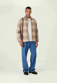 Beige plaid shacket over a white t-shirt, paired with blue jeans featuring a patch pocket, and black shoes. Model stands against a light background.