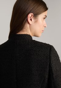 Zwarte textuurjacket met een hoge kraag, voorzien van een geweven patroon en subtiele details, weergegeven vanuit de achterkant, waarbij de snit en stof worden benadrukt.