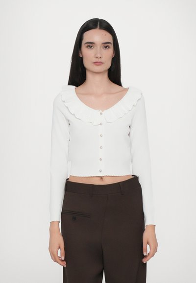 Cardigan blanc court avec un col ondulé à bords festonnés, des manches longues et des fermetures à boutons agrémentées de détails décoratifs.