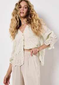 Ein Model mit welligem, langem Haar trägt eine leichte, strukturierte Strickjacke und lockere beige Hosen. Sie posiert mit einer Hand auf der Hüfte vor einem neutralen Hintergrund.