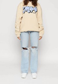Beżowy hoodie z niebieskim graficznym tekstem, duża przednia kieszeń. Jasnoniebieskie jeansy z przetarciami na kolanach, w połączeniu z białymi trampkami.