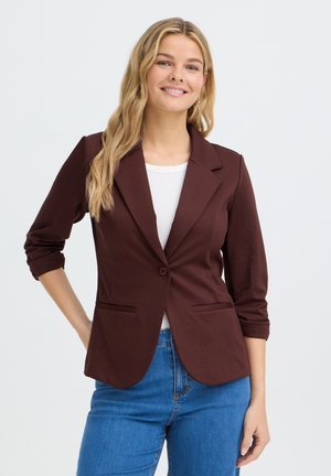 Femme aux longs cheveux blonds portant un blazer marron sur un haut blanc et un jean bleu, souriante, une main sur la hanche.