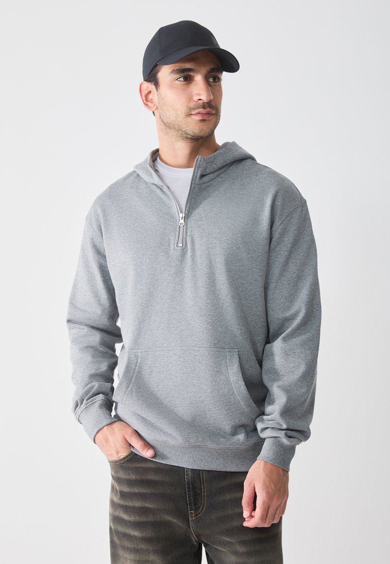 Grå hettegenser med glidelås i halsen, frontlomme og ribbestrikkede mansjetter, båret over en hvit t-skjorte, sammen med mørke jeans.